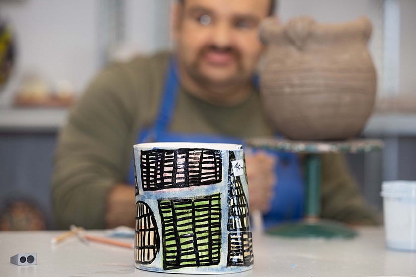A painted ceramic mug in focus in the foreground, with a pot on a stand and a person behind out of focus in the background.